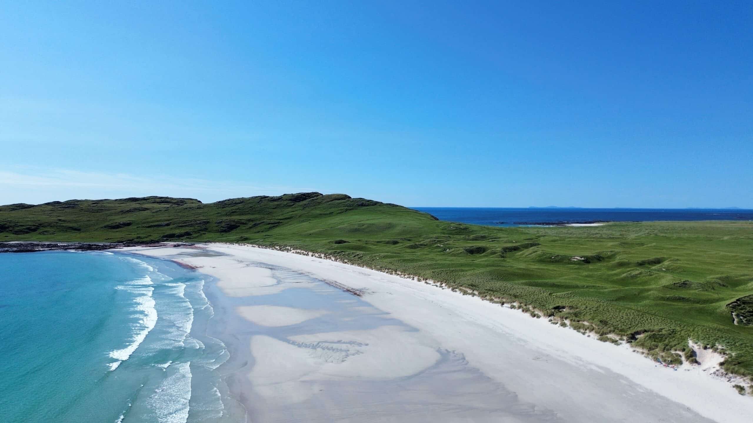 Serene coastal landscape with sandy beach, rolling green hills, and bright blue sky.