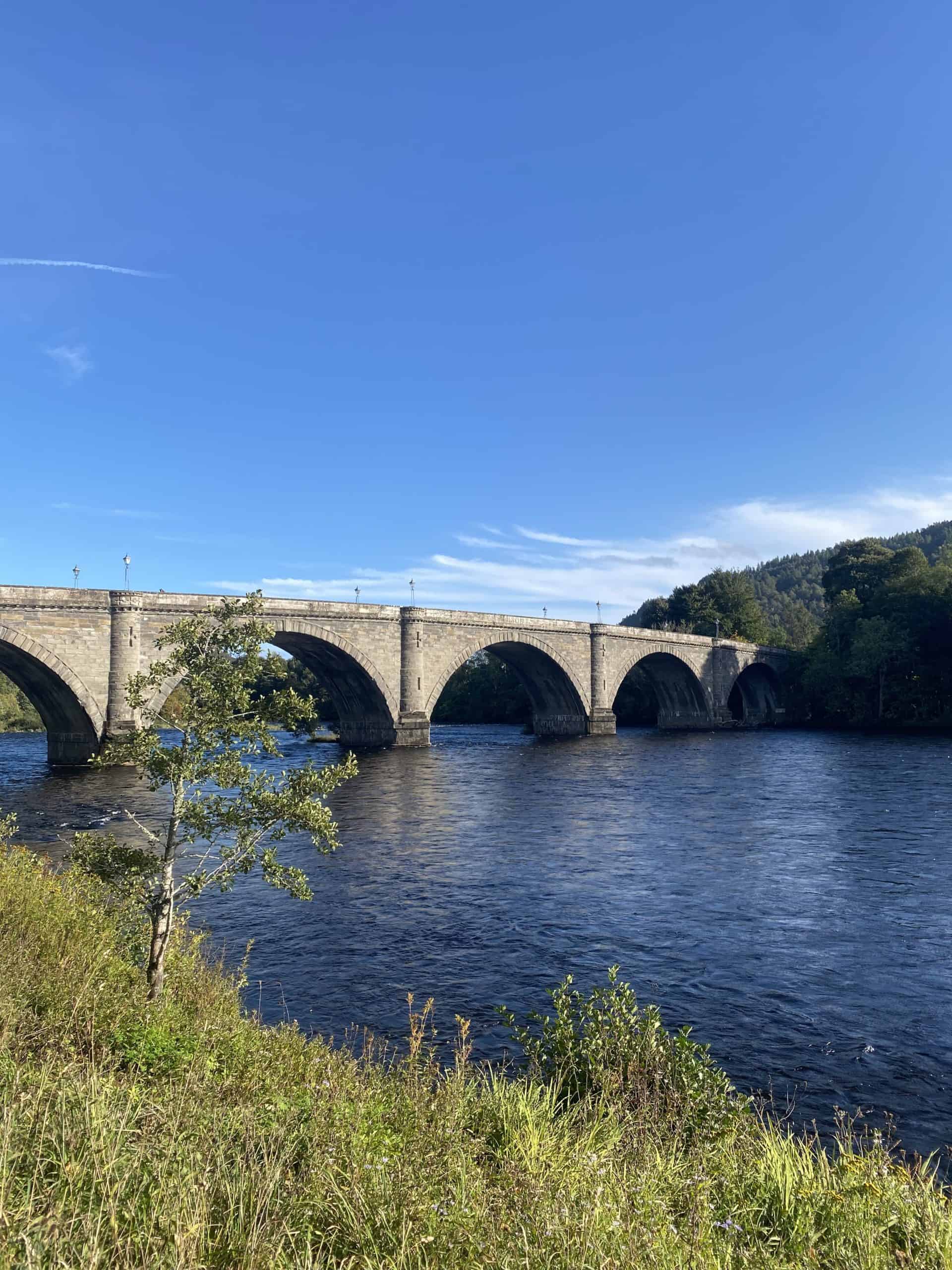 Stone railway bridge over river with lush greenery and blue sky in the background, scenic outdoor landscape, Wee Trails.