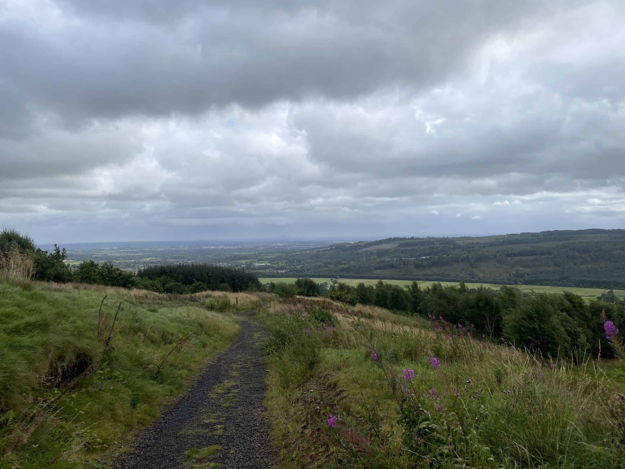 Winding gravel trail through lush green countryside with rolling hills and cloudy sky.