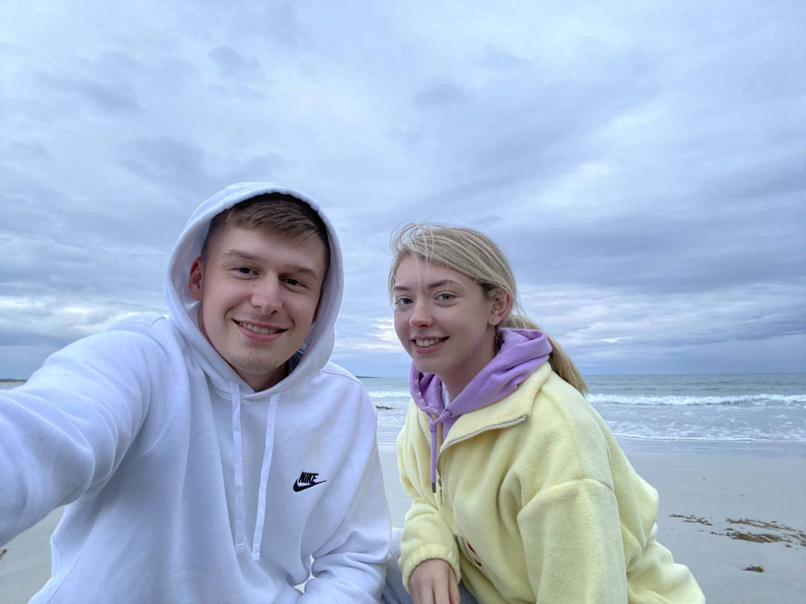 Smiling teenagers taking a selfie on the beach with cloudy sky and ocean background.
